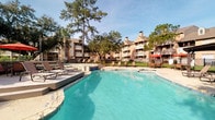 This community's amenities center on a spacious outdoor pool that serves as a central gathering point, surrounded by a generous sun deck outfitted with lounge chairs and framed by mature trees. The pool's clear turquoise water and stone-accented edges lend a resort-like atmosphere, while the multi-story residential buildings visible in the background reinforce the sense of a well-established community setting.

The leasing office is organized across a single floor and opens into a comfortable lounge area featuring vaulted ceilings, wood-look flooring, and a ceiling fan overhead. Warm accent colors — vibrant orange and teal — are carried throughout via throw pillows, an area rug, and drapery panels flanking tall windows that draw in natural light and views of the surrounding trees. A wall-mounted TV, glass coffee table, and seating arrangement create an inviting, relaxed environment for visitors.

Adjacent to the lounge, a private office workspace offers a substantial wood desk, upholstered guest chairs, and multiple windows that continue the theme of abundant natural light. A hallway connecting the spaces features wood-look flooring, built-in closet storage, and tile flooring transitions, offering a functional and polished passageway throughout the office.