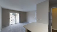 This single-floor space features an open-concept layout that seamlessly connects the kitchen and living areas. The living room is finished with wall-to-wall carpeting in a neutral gray tone and painted in warm greige, creating a cohesive and understated aesthetic throughout. A sliding glass door along one wall floods the space with natural light and provides direct access to an outdoor balcony, where a white railing is visible along with views of neighboring buildings and mature trees, suggesting a peaceful residential setting.

The kitchen area is partially visible in the foreground, highlighted by a light-toned countertop with subtle speckled detailing, offering a functional surface that extends into the living space. A white interior door is positioned along the far wall, providing access to additional rooms. The smooth white ceiling maintains a clean, uniform appearance overhead. Electrical outlets and light switch plates are evenly distributed along the walls. The overall design is practical and move-in ready, with a neutral palette that offers flexibility for personalization.