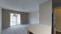 This single-floor space features an open-concept layout that seamlessly connects the kitchen and living areas. The living room is finished with wall-to-wall carpeting in a neutral gray tone and painted in warm greige, creating a cohesive and understated aesthetic throughout. A sliding glass door along one wall floods the space with natural light and provides direct access to an outdoor balcony, where a white railing is visible along with views of neighboring buildings and mature trees, suggesting a peaceful residential setting.

The kitchen area is partially visible in the foreground, highlighted by a light-toned countertop with subtle speckled detailing, offering a functional surface that extends into the living space. A white interior door is positioned along the far wall, providing access to additional rooms. The smooth white ceiling maintains a clean, uniform appearance overhead. Electrical outlets and light switch plates are evenly distributed along the walls. The overall design is practical and move-in ready, with a neutral palette that offers flexibility for personalization.