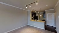 Located at 2015 Copperleaf Pkwy in Durham, North Carolina, this single-floor residence offers a thoughtfully designed open-concept layout that seamlessly connects the living and kitchen areas. The living space features neutral-toned carpet flooring and soft grey walls accented by crisp white crown molding, creating a clean and welcoming atmosphere throughout.

The kitchen is a standout feature, appointed with dark cabinetry that provides a rich contrast against the warm-toned breakfast bar countertop. A stainless steel sink is integrated into the bar-height peninsula, which serves as both a functional workspace and a casual dining area. Pendant lighting hangs above the peninsula, complementing the additional track lighting that runs across the ceiling to illuminate the kitchen and living space effectively. Stainless steel appliances, including a refrigerator and range, complete the kitchen's modern aesthetic.

Track lighting is a recurring design element throughout the space, providing flexible, directed illumination across the main living area. The open floor plan allows for a natural flow between spaces, making the residence feel airy and well-connected. Crown molding detailing adds a refined architectural touch that elevates the overall finish of the interior. This residence offers a practical yet stylish single-level living experience in Durham, NC.