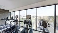 This contemporary fitness center is designed to energize and inspire, combining a well-appointed exercise environment with an abundance of natural light. The defining architectural feature is the expansive floor-to-ceiling window wall that spans the length of the space, offering panoramic views of the surrounding urban landscape and filling the room with daylight throughout the day. The elevated vantage point provides an engaging backdrop of nearby buildings, parking areas, and open sky, making workouts feel open and invigorating rather than confined. The flooring features a patterned, commercial-grade carpet designed to withstand heavy foot traffic while adding a modern aesthetic. The equipment layout is thoughtfully arranged to take full advantage of the window views, with a row of premium cardio machines — including True-brand elliptical cross-trainers and treadmills — positioned directly facing the glass. The clean white ceiling and bright, neutral interior palette keep the space feeling fresh and uncluttered. Overall, the fitness center strikes a balance between function and comfort, offering a bright, well-equipped environment suited for a focused and enjoyable workout experience.