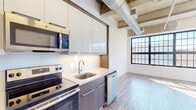 Located at 121 Westmoreland Ave in White Plains, NY, this single-floor unit showcases a compelling blend of industrial character and contemporary finishes. The space is defined by its soaring exposed ceilings, where raw concrete beams, visible ductwork, and linear fluorescent lighting contribute an authentic loft-like aesthetic throughout.

Large, multi-pane factory-style windows are a standout architectural feature, framing views of the surrounding White Plains landscape and flooding the interior with generous natural light. Wood-look plank flooring in a light, neutral tone runs consistently through the main living areas, complementing the industrial overhead elements with a warm, modern contrast.

The kitchen is well-appointed with stainless steel appliances—including a range, microwave, and dishwasher—paired with sleek, high-gloss upper cabinetry and wood-toned lower cabinets set against a clean white backsplash. A bedroom area features a sliding door closet and a prominent window with outdoor views. The bathroom is finished with large-format tile flooring and neutral gray walls, providing a clean, functional space. A louvered bi-fold door adds a subtle design detail near the kitchen entry. Overall, the unit offers a distinctive, light-filled environment with strong industrial character.