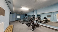 Located at 2210 Sullivan Road in College Park, Georgia, this single-floor fitness facility offers a comprehensive workout environment spread across a generous footprint. The space is divided into two distinct zones, each designed to support a different style of training.

The cardio and functional training area features light blue walls, textured ceilings with fluorescent lighting, and durable gray carpet flooring. A row of True-brand treadmills, ellipticals, and stationary bikes lines the room, complemented by full-length wall mirrors that amplify the sense of space. A wall-mounted television provides entertainment during workouts, and a staircase area connects to additional access points within the facility.

The strength and functional training zone transitions to warm hardwood-style flooring and benefits from recessed skylights that brighten the room with overhead illumination. This area is stocked with a multi-station cable machine, adjustable benches, a comprehensive dumbbell rack, medicine balls, kettlebells, battle ropes, a heavy tire, and TRX suspension trainers — catering to a wide range of functional and strength-based workouts. A separate spin studio area features stationary cycling bikes on hardwood flooring with mirrored walls, ideal for group cycling sessions. Throughout, the consistent blue color palette and clean layout create an energizing and focused training atmosphere.