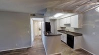 This single-floor residence offers a practical and well-organized layout with a clean, contemporary aesthetic. The kitchen serves as the heart of the home, featuring crisp white cabinetry that provides ample storage, paired with dark countertops that offer a striking visual contrast. Black appliances, including a refrigerator, dishwasher, and cooktop, complement the modern color palette. A central kitchen island adds additional prep space and serves as a natural dividing point between the kitchen and the adjoining living areas. A skylight box above the kitchen fills the space with natural light, enhancing the overall brightness of the interior. Wood-style plank flooring runs consistently throughout, lending warmth and cohesion to the space. The textured ceiling and neutral gray walls create a subtle, understated backdrop that allows the kitchen's design elements to stand out. A hallway visible from the kitchen leads to additional rooms, suggesting a thoughtful and functional floor plan. The HVAC vent integrated into the ceiling ensures year-round comfort. Overall, this residence balances style and practicality in a cohesive, move-in-ready package.