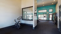 This single-floor amenity space is divided into two distinct functional areas: a fitness center and a laundry facility. The fitness center is the standout feature, boasting impressively high ceilings that create an open, airy atmosphere. Bold teal accent walls add a vibrant, energizing touch, while large grid-paned windows and glass doors flood the space with natural light and provide views of the surrounding outdoor area, including a glimpse of a pool. The dark carpeted flooring provides a practical, cushioned surface throughout the workout area. Equipment includes treadmills, stationary and spin bikes, a rowing machine, strength training cable machines, and additional cardio equipment, all complemented by wall-to-wall mirrors that visually expand the space. Elegant black-framed French doors with glass panels connect the fitness areas, adding a refined architectural detail. The adjacent laundry facility features stacked washer and dryer units, tile flooring in a classic checkerboard pattern, a central folding counter, and windows that bring in natural light. The teal accent walls continue into this area, maintaining a cohesive visual identity throughout the entire space. Together, both areas offer practical, well-designed amenities in a bright and welcoming environment.