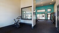 This single-floor amenity space is divided into two distinct functional areas: a fitness center and a laundry facility. The fitness center is the standout feature, boasting impressively high ceilings that create an open, airy atmosphere. Bold teal accent walls add a vibrant, energizing touch, while large grid-paned windows and glass doors flood the space with natural light and provide views of the surrounding outdoor area, including a glimpse of a pool. The dark carpeted flooring provides a practical, cushioned surface throughout the workout area. Equipment includes treadmills, stationary and spin bikes, a rowing machine, strength training cable machines, and additional cardio equipment, all complemented by wall-to-wall mirrors that visually expand the space. Elegant black-framed French doors with glass panels connect the fitness areas, adding a refined architectural detail. The adjacent laundry facility features stacked washer and dryer units, tile flooring in a classic checkerboard pattern, a central folding counter, and windows that bring in natural light. The teal accent walls continue into this area, maintaining a cohesive visual identity throughout the entire space. Together, both areas offer practical, well-designed amenities in a bright and welcoming environment.
