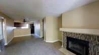 This single-floor space presents a straightforward and functional layout designed for comfortable everyday living. The living area is carpeted and anchored by a notable fireplace finished with a mosaic tile surround and a clean white mantel, serving as a natural focal point of the room. Warm beige walls carry throughout the space, creating a neutral and cohesive atmosphere.

The open sightlines from the living area extend toward a hallway and adjacent kitchen zone, which is partially visible and features dark appliances, including a refrigerator, along with a ceiling fan that aids in ventilation and comfort. The transition between spaces flows naturally, with the hallway offering access to additional rooms or storage, indicated by a paneled door along the corridor.

White baseboards provide clean contrast against the neutral walls and carpeted flooring. The layout makes efficient use of the available space, with no wasted areas. Overall, the space maintains a simple, move-in-ready aesthetic with practical features, including the fireplace as a standout architectural element that adds character to an otherwise understated interior.