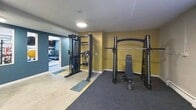 Located at 5100 Leetsdale Drive in Denver, Colorado, this 2,480-square-foot amenity floor offers residents a well-rounded collection of shared spaces designed for comfort, community, and fitness. The welcoming entrance hallway features wood-look flooring and a modern package locker system for secure parcel retrieval. Adjacent to the entry, a stylish living room serves as the social hub of the space, furnished in a bold mid-century modern aesthetic with vibrant teal, orange, and yellow upholstered seating, a wood coffee table, patterned area rugs, and retro artwork adorning the walls. A large interior window connects the lounge to the broader common area, enhancing the open feel. The fitness amenities are divided across two dedicated exercise rooms. The first houses cardio equipment including treadmills and an elliptical trainer, with wall-mounted televisions and windows providing natural light. The second exercise room is equipped with strength training machines, including a cable station and a power rack with a bench, set against bold teal and mustard-yellow accent walls. The shared bathroom features eye-catching multicolored mosaic tile work, a wall-mounted sink with gold fixtures, an oval mirror, and accessibility grab bars, combining practicality with vibrant design.