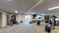 Located at 2770 E Uintah St in Colorado Springs, Colorado, this single-floor fitness center offers a well-equipped workout space designed to support a variety of exercise routines. The room features textured ceilings with flush skylight-style panels that provide bright, even lighting throughout. Durable, low-pile carpet tiles cover the floors, offering a practical and comfortable surface underfoot. A full wall of floor-to-ceiling mirrors lines one side of the space, visually expanding the room and allowing users to monitor their form during workouts. Cardio equipment includes a treadmill, stationary bike, and elliptical trainer, while a multi-function cable weight machine and adjustable bench cater to strength training needs. Exercise accessories such as stability balls are also available. A wall-mounted flat-screen television adds entertainment during workouts. Natural light filters in through a glass sliding door that opens to an outdoor area, lending the space an airy, open feel. A secondary area within the fitness center houses an additional cardio machine, a storage shelving unit, and posted informational materials, along with a floor fan for added ventilation. A Coca-Cola vending machine provides convenient access to beverages. The overall ambiance is clean, functional, and well-maintained, making it a practical facility for everyday fitness needs.