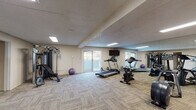 Located at 2770 E Uintah St in Colorado Springs, Colorado, this single-floor fitness center offers a well-equipped workout space designed to support a variety of exercise routines. The room features textured ceilings with flush skylight-style panels that provide bright, even lighting throughout. Durable, low-pile carpet tiles cover the floors, offering a practical and comfortable surface underfoot. A full wall of floor-to-ceiling mirrors lines one side of the space, visually expanding the room and allowing users to monitor their form during workouts. Cardio equipment includes a treadmill, stationary bike, and elliptical trainer, while a multi-function cable weight machine and adjustable bench cater to strength training needs. Exercise accessories such as stability balls are also available. A wall-mounted flat-screen television adds entertainment during workouts. Natural light filters in through a glass sliding door that opens to an outdoor area, lending the space an airy, open feel. A secondary area within the fitness center houses an additional cardio machine, a storage shelving unit, and posted informational materials, along with a floor fan for added ventilation. A Coca-Cola vending machine provides convenient access to beverages. The overall ambiance is clean, functional, and well-maintained, making it a practical facility for everyday fitness needs.