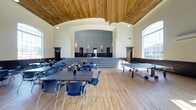 This recreation center is a thoughtfully designed, single-floor facility that combines classic architectural charm with functional, multi-use space. The centerpiece of the building is its expansive main hall, which immediately draws the eye upward to a stunning vaulted ceiling clad in warm, natural wood — a standout architectural detail that adds character and grandeur to the room. The arched ceiling transitions seamlessly into crisp white walls, creating a bright and welcoming ambiance throughout.

The hall features rich hardwood flooring that extends across the entire space, lending warmth underfoot while remaining durable for heavy use. A raised stage at the far end of the room is framed by a wide, decorative archway with ornate molding, making it well-suited for performances, presentations, or special events. Steps lead up to the stage, which opens into an additional room beyond, offering flexible overflow or backstage space.

The main hall is outfitted with long rectangular tables and blue chairs arranged for group gatherings, and a ping-pong table along one side speaks to the space's recreational purpose. Large windows along the side wall invite generous natural light, enhancing the open and airy feel of the interior. Pendant lighting supplements the natural light, ensuring the space is well-lit at all hours. Overall, this recreation center offers a welcoming, versatile environment suited to a wide range of community activities.