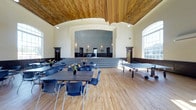This recreation center is a thoughtfully designed, single-floor facility that combines classic architectural charm with functional, multi-use space. The centerpiece of the building is its expansive main hall, which immediately draws the eye upward to a stunning vaulted ceiling clad in warm, natural wood — a standout architectural detail that adds character and grandeur to the room. The arched ceiling transitions seamlessly into crisp white walls, creating a bright and welcoming ambiance throughout.

The hall features rich hardwood flooring that extends across the entire space, lending warmth underfoot while remaining durable for heavy use. A raised stage at the far end of the room is framed by a wide, decorative archway with ornate molding, making it well-suited for performances, presentations, or special events. Steps lead up to the stage, which opens into an additional room beyond, offering flexible overflow or backstage space.

The main hall is outfitted with long rectangular tables and blue chairs arranged for group gatherings, and a ping-pong table along one side speaks to the space's recreational purpose. Large windows along the side wall invite generous natural light, enhancing the open and airy feel of the interior. Pendant lighting supplements the natural light, ensuring the space is well-lit at all hours. Overall, this recreation center offers a welcoming, versatile environment suited to a wide range of community activities.
