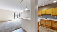 This single-floor unit is thoughtfully designed with an open, connected layout that promotes a sense of spaciousness. The kitchen and living area share a semi-open floor plan, separated by a half-wall partition topped with a breakfast bar ledge. Pendant lights hang above this dividing wall, while track lighting runs throughout, providing flexible illumination across the space.

The kitchen features warm-toned oak cabinetry with ample upper and lower storage, white appliances including a refrigerator, range, and dishwasher, and a double sink. Stone-patterned tile flooring extends through the kitchen and into the bathroom, offering a cohesive, durable surface in these functional areas.

The living area is generously proportioned, carpeted in neutral grey tones, and benefits from large windows that bring in natural light and views of the surrounding exterior. Additional carpeted rooms serve as bedrooms, each with oversized windows that flood the spaces with sunlight.

The bathroom is designed with accessibility in mind, featuring multiple grab bars positioned near the toilet and throughout the room, a wood-toned vanity with a wide mirror, and the same tile flooring found in the kitchen. The overall color palette is neutral and understated, creating a calm, functional atmosphere throughout.