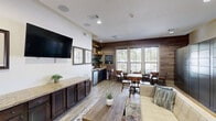 Located at 5350 Old Dowlen Rd in Beaumont, TX, this single-floor community lounge space offers a welcoming and well-appointed environment designed for comfort and convenience. The living area features light wood-style flooring, a tufted cream sofa with blue accent pillows, a rustic wood coffee table, and a colorful area rug that anchors the seating arrangement. A wall-mounted flat-screen TV is flanked by a gallery of framed black-and-white portraits, while dark espresso cabinetry with granite countertops lines the adjacent wall, adding a refined, upscale character to the room. Natural light streams through multiple large windows dressed with blinds, offering views of the surrounding greenery and brightening the entire space. A dining area with wooden tables and chairs is positioned near the windows, creating an inviting spot for casual gatherings. The kitchenette is outfitted with mosaic tile backsplash, open wood shelving stocked with mugs and a coffee maker, a compact refrigerator, and granite countertops — ideal for a self-serve coffee station. A dedicated package locker wall with electronic access provides practical functionality. Recessed lighting and in-ceiling speakers complete the polished, modern-rustic ambiance throughout.