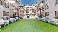Located at 3234 North Scottsdale Road in Scottsdale, Arizona, this beautifully designed outdoor courtyard serves as a central gathering hub within a contemporary multi-story residential community. The space is defined by a sweeping expanse of vibrant, manicured green lawn that anchors the entire courtyard, creating an inviting and open atmosphere beneath the Arizona sky.

A striking red steel pergola structure serves as the architectural centerpiece, its angular, intersecting beams casting dramatic shadows across the space and adding a bold, artistic flair to the outdoor environment. Surrounding the lawn, multiple seating vignettes are thoughtfully arranged, featuring woven lounge chairs and cushioned sofas with navy blue accents, ideal for relaxed socializing or quiet outdoor enjoyment.

A central fire pit seating area provides an inviting focal point for evening gatherings, encircled by modern curved lounge chairs. Mature palm trees and carefully maintained desert-friendly landscaping add natural texture and shade throughout the courtyard.

The space is fully enclosed by the modern residential building, whose multi-level balconies, large windows, and clean stucco facade overlook the courtyard from all sides, creating a sense of privacy and community. Additional amenities such as outdoor grilling stations are integrated into the design, making this courtyard a comprehensive and resort-inspired outdoor living experience.