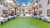 Located at 3234 North Scottsdale Road in Scottsdale, Arizona, this beautifully designed outdoor courtyard serves as a central gathering hub within a contemporary multi-story residential community. The space is defined by a sweeping expanse of vibrant, manicured green lawn that anchors the entire courtyard, creating an inviting and open atmosphere beneath the Arizona sky.

A striking red steel pergola structure serves as the architectural centerpiece, its angular, intersecting beams casting dramatic shadows across the space and adding a bold, artistic flair to the outdoor environment. Surrounding the lawn, multiple seating vignettes are thoughtfully arranged, featuring woven lounge chairs and cushioned sofas with navy blue accents, ideal for relaxed socializing or quiet outdoor enjoyment.

A central fire pit seating area provides an inviting focal point for evening gatherings, encircled by modern curved lounge chairs. Mature palm trees and carefully maintained desert-friendly landscaping add natural texture and shade throughout the courtyard.

The space is fully enclosed by the modern residential building, whose multi-level balconies, large windows, and clean stucco facade overlook the courtyard from all sides, creating a sense of privacy and community. Additional amenities such as outdoor grilling stations are integrated into the design, making this courtyard a comprehensive and resort-inspired outdoor living experience.