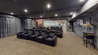 This single-floor entertainment space is purpose-built for a premium theater experience. The main screening area is anchored by multiple rows of black leather recliner-style seats with built-in cup holders, arranged in a tiered layout ideal for unobstructed viewing. The ceiling is finished in a deep charcoal tone with recessed lighting that creates a dramatic, cinema-like ambiance, while dark wood beam accents add architectural definition.

Adjacent to the seating area, a dedicated lobby or foyer space features gray-painted walls, neutral striped carpet tiles, and a classic popcorn cart — reinforcing the movie-house theme. A dark wood-framed doorway connects this entry zone to the main hall.

Beyond the theater seating, an open lounge area transitions with lighter wall tones, arched window details framed by drapery, a decorative chandelier, and framed artwork, providing a welcoming space for gathering before or after screenings. Large double entry doors with glass panels allow access from a corridor, and exit signage confirms the space meets safety standards. The overall design balances functional entertainment infrastructure with refined decorative touches throughout.