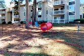 This outdoor dog park is a dedicated pet amenity situated within a multi-building residential community, enclosed by a black chain-link fence with a gated entry point for controlled access. The space is set at ground level and features a natural grass and dirt surface shaded by a canopy of mature pine and deciduous trees, creating a comfortable environment for pets year-round. Colorful agility equipment is distributed throughout the area, including a red tunnel, a blue slide-style obstacle, and a blue ring station, offering interactive enrichment opportunities for dogs of varying sizes. The park is clearly divided into sections, suggesting separate areas that may accommodate different dog sizes. Surrounding the perimeter, views of the multi-story residential buildings are visible, along with paved parking areas and maintained walkways. The grounds are bathed in natural sunlight filtered through the tree canopy, lending a lively, open-air atmosphere. Waste disposal stations are positioned within the space to promote cleanliness and responsible pet ownership. The overall setting is practical and well-integrated into the broader community landscape, providing residents and their pets a safe and engaging outdoor retreat.