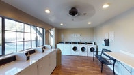 The laundry room at 2660 Alpine is a well-appointed shared facility designed with both functionality and comfort in mind. The space features warm hardwood-style flooring that runs throughout, lending a clean and welcoming atmosphere uncommon in utility spaces of this kind. A row of front-loading washing machines lines the back wall, accompanied by a set of top-loading washers and dryers along the side, providing ample capacity for residents. Recessed lighting in the ceiling ensures the room is well-illuminated, while a ceiling fan with a light fixture adds air circulation and additional brightness. Large windows along one wall allow natural light to filter in, creating an open and airy feel. A payment kiosk is conveniently located for easy access. A decorative metal bench is positioned near the entrance, offering a comfortable spot to wait. The neutral-toned walls and tidy layout contribute to a space that is both practical and pleasant to use. Overall, this laundry facility is thoughtfully designed to serve its residents efficiently while maintaining a clean, comfortable environment.