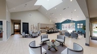 This elegantly designed single-floor community space offers an open, airy atmosphere defined by dramatic vaulted ceilings that soar overhead, flooding the interior with natural light through strategically placed skylights. Light-toned tile flooring runs throughout, complementing the neutral color palette accented by a bold teal feature wall that adds a striking visual anchor to the space.

The main gathering area is thoughtfully arranged with multiple distinct seating vignettes, each furnished with stylish upholstered chairs, glass-topped coffee tables, and plush area rugs. Decorative chandeliers hang from the high ceilings, adding a refined elegance. A media lounge area features a large wall-mounted television, built-in cabinetry, and comfortable seating designed for relaxed viewing.

The fully equipped kitchen is a standout feature, showcasing floor-to-ceiling gray shaker cabinetry, a textured tile backsplash, stainless steel appliances, and a generous dark stone-topped island with upholstered bar seating. This space transitions seamlessly into a dining area, making it ideal for entertaining and gatherings.

A corridor with glass-panel partitions connects additional private rooms, while large windows along one wall offer views of an outdoor pool area, bringing the outdoors in. The overall ambiance is modern, welcoming, and polished — a premium space designed for both social and relaxed use.