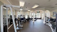 This fitness center offers a generously sized, single-floor workout space designed to accommodate a full range of exercise needs. The room's architectural character is defined by elegant arched windows that line the walls, allowing abundant natural light to flood the interior and provide views of the surrounding greenery. Decorative white columns punctuate the space, adding a classic touch while visually dividing distinct workout zones.

The flooring throughout is durable rubber matting, practical for heavy equipment use and providing traction and cushioning underfoot. Large wall-mounted mirrors expand the sense of space and allow users to monitor their form during exercise. Recessed and panel ceiling lights supplement the natural light, ensuring the space remains well-illuminated at all times.

The cardio area features Cybex treadmills, elliptical trainers, stationary bikes, and a rowing machine, all positioned near the windows to make use of natural light and outdoor views. The strength-training section is well-stocked with cable machines, multi-functional weight stations, benches, and free-weight racks. A wall-mounted flat-screen television is positioned for easy viewing from the cardio zone.

The two-tone wall treatment — warm beige above and deep navy below — gives the space a polished, purposeful aesthetic, reinforcing its identity as a dedicated, professionally appointed fitness facility.