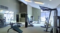 This fitness center is a thoughtfully designed exercise facility that combines functionality with an open, airy atmosphere. The space features notably high, angled ceilings that amplify the sense of volume and contribute to an energizing workout environment. Large floor-to-ceiling windows line one side of the room, flooding the interior with abundant natural light and offering views of a snowy outdoor landscape, creating a refreshing connection to the outdoors.

The facility is outfitted with a comprehensive selection of exercise equipment, including a multi-function cable/weight machine, an adjustable bench, a leg press or seated strength machine, an elliptical trainer, and a stationary bike, among other cardio and resistance options. The equipment is arranged to allow comfortable circulation throughout the space. Neutral-toned walls in a muted green-grey palette, combined with light-colored flooring, create a clean and modern aesthetic. Recessed and panel lighting supplements the natural light, ensuring the space remains well-lit regardless of the time of day. A mirrored wall section enhances the sense of depth and supports proper form during workouts. An additional enclosed room or office area is visible at the rear of the space, suggesting potential for private training or administrative use.