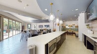 This single-floor clubhouse is designed as a versatile communal gathering space, blending contemporary aesthetics with functional amenities. The layout flows seamlessly across several distinct zones, unified by light stone-toned tile flooring and a consistent slate-blue ceiling treatment that adds depth and visual interest throughout.

At the heart of the space is a large central kitchen island topped with bright white countertops, illuminated by a striking trio of teardrop-shaped glass pendant lights with brass fixtures. Adjacent cabinetry in dark navy features a herringbone-patterned tile backsplash, a sink, and built-in appliances, with mounted flat-screen TVs providing an engaging focal point. Bar-height seating lines one side of the island, creating an inviting social hub.

The lounge area offers a comfortable arrangement of upholstered chairs and sofas, anchored by a sculptural wood-and-metal media console beneath two wall-mounted screens. A banquette with bold geometric-patterned upholstery adds a pop of color nearby.

A private conference room accommodates a seated group around a dark wood table, decorated with vibrant artwork and benefiting from large sliding glass doors that open to an outdoor courtyard, filling the room with natural light and outdoor views.