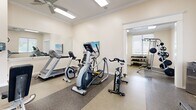 Located at 3895 Waterside Dr in Elizabeth City, North Carolina, this fitness center is a well-equipped, single-room exercise facility designed to support a well-rounded workout routine. The space features a durable speckled composite floor with contrasting mat zones that delineate cardio and strength training areas, contributing to both functionality and visual organization.

The cardio section is outfitted with Matrix-brand equipment, including a treadmill, elliptical trainer, and stationary cycle, providing a variety of aerobic exercise options. A large wall-mounted mirror reflects the space and enhances the sense of openness. A flat-screen television is mounted on the wall, offering entertainment during workouts.

The strength training area features a multi-functional cable and weight machine loaded with plate weights, a flat workout bench, and a full dumbbell rack with a wide range of free weights. Natural light filters into the room through multiple windows fitted with horizontal blinds, and a glass-paneled exterior door opens to an outdoor view, lending an airy quality to the interior.

Crown molding runs along the ceiling perimeter, and ceiling fans equipped with overhead fluorescent lighting ensure consistent illumination and airflow. Decorative fish wall sculptures and potted plants add a welcoming, coastal-inspired touch that reflects the character of the Elizabeth City area.