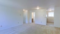 This single-floor residential space offers a clean, functional layout designed for comfortable everyday living. The interior is characterized by light-toned wall-to-wall carpeting that adds warmth and softness underfoot throughout the main living areas. Smooth white walls and a textured white ceiling contribute to a bright, airy atmosphere that feels open and welcoming.

The space features flush-mount ceiling light fixtures that provide consistent ambient lighting across the rooms. A doorway visible from the main room leads into a connected secondary space, which appears to contain cabinetry — likely a bathroom or walk-in closet area — offering practical storage and utility within close proximity to the primary living space. A secondary door on the same wall provides additional access or closet storage.

Electrical outlets and switch plates are neatly positioned along the walls, reflecting a thoughtfully planned interior. The overall aesthetic is neutral and move-in ready, with a simple, understated design that allows for flexible personalization. The single-floor layout ensures easy navigation throughout, making the space accessible and efficient. This property presents a well-maintained, straightforward residential environment suited for a range of occupants.