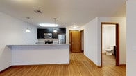 This single-floor space offers a clean, modern aesthetic with an open and functional layout. The kitchen serves as a central highlight, featuring dark cabinetry that contrasts beautifully with light granite countertops and a full suite of stainless steel appliances, including a refrigerator, microwave, and electric range. A peninsula countertop extends the workspace and provides an inviting area for casual dining or meal preparation. Pendant lighting above the kitchen area adds a warm, focused glow, complementing the flush ceiling fixtures throughout the space. Warm-toned hardwood flooring flows seamlessly across the entire unit, lending a cohesive and inviting feel to every area. The open layout connects the kitchen to the living area, creating a spacious and airy atmosphere ideal for both everyday living and entertaining. A solid wood entry door with warm tones provides a welcoming transition from the exterior. A glimpse of an adjacent room — likely a bathroom — is visible at the far end of the hallway, suggesting a thoughtfully organized floor plan. The overall ambiance is bright, contemporary, and well-appointed, making efficient use of the available space.