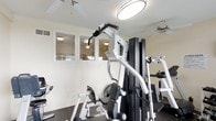 This fitness center is designed to support a well-rounded workout routine within a clean, functional environment. The space is anchored by a large, multi-station cable and weight machine in a classic black-and-white finish, offering a variety of strength training exercises from a single unit. Additional cardio equipment is visible throughout the room, providing options for a comprehensive exercise session. The flooring is dark-toned, offering a practical and durable surface suited for physical activity. Overhead, circular flush-mount light fixtures provide bright, even illumination, while multiple ceiling fans ensure consistent airflow and comfort during workouts. Natural light filters in through a row of horizontal-slatted windows along one wall, contributing to an open and inviting atmosphere. The walls are finished in a neutral, light tone, keeping the space feeling clean and uncluttered. Mirrors are positioned to allow users to monitor their form during exercise. The overall ambiance is practical and purposeful, with every element oriented toward creating an efficient and comfortable fitness environment.