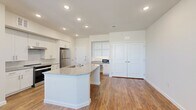 This single-floor, one-bedroom, one-bathroom unit offers a thoughtfully designed open-concept layout that maximizes functionality and flow. The kitchen serves as the centerpiece of the living area, showcasing white shaker cabinetry, granite countertops, a large central island with a built-in sink, and a full suite of stainless steel appliances including a refrigerator and range. Recessed lighting illuminates the space throughout, complementing the warm hardwood-style flooring that runs across the main living areas. A built-in desk nook with open shelving and granite countertop provides a dedicated workspace seamlessly integrated into the kitchen area, while a set of double doors leads to additional storage. The bedroom features plush carpeting, a ceiling fan, and dual windows that allow natural light and outdoor views. It connects to a walk-in closet with multi-tiered shelving and carpet flooring. The bathroom is finished with granite countertops, white cabinetry, a large mirror with vanity lighting, and a tiled shower-tub combination equipped with grab bars and a handheld showerhead. Hardwood-style flooring extends into the bathroom, maintaining a cohesive aesthetic throughout the unit.