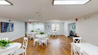 This single-floor facility is thoughtfully designed to provide comfortable, functional communal spaces with a clean and inviting aesthetic. The centerpiece of the space is a generously sized dining and activity room featuring rich, warm-toned hardwood flooring that extends throughout the area. The room is outfitted with multiple round white tables and matching cross-back chairs, arranged to encourage social interaction. Fresh green plants serve as cheerful centerpieces, adding life to the bright, light-gray painted walls accented by classic chair rail wainscoting. Overhead, flush-mounted fluorescent lighting ensures the room is well-lit, while a window allows natural light to filter in. Wall art depicting nature scenes adds a calm, decorative touch, and a beverage station is conveniently positioned along one wall. Adjacent to the dining area is a relaxed lounge space, carpeted in a soft gray tone and furnished with plush blue sofas, coordinating accent pillows, and a cottage-style coffee table. Ceiling fans provide comfortable air circulation, and large windows with shutters bring in natural light. A bookshelf and tasteful artwork complete the lounge's welcoming atmosphere. Together, these spaces reflect a facility designed with residents' comfort and community in mind.