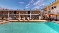 This outdoor pool area in Lafayette, Louisiana, is a well-appointed communal amenity designed for relaxation and recreation. The centerpiece is a generously sized swimming pool with clear turquoise water, framed by a classic brick paver deck that extends along its perimeter. A covered patio runs along one side of the pool, supported by white-painted structural posts and featuring a painted panel ceiling, providing a shaded retreat from the Louisiana sun. This covered area includes direct access to an interior room with pool rules posted prominently, indicating organized management of the space. The pool deck is furnished with modern lounge chairs and a gas grill, creating a comfortable outdoor living environment. The area is fully enclosed by a black iron security fence, ensuring safety and privacy. Mature trees are visible beyond the fence line, adding a natural backdrop. The surrounding two-story brick building features open walkways, iron railings, and staircases, contributing to the classic architectural character of the property. Large sliding glass doors from ground-floor units open directly onto the pool area, offering immediate outdoor access and unobstructed pool views from within.