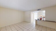 This single-floor unit offers a clean, functional layout with a light and airy feel throughout. The living area is finished with large-format cream ceramic tile flooring, neutral cream-toned walls, and a textured ceiling, creating a bright and welcoming atmosphere. A window fitted with vertical blinds allows natural light to fill the space, while a front entry door provides convenient access.

The kitchen is seamlessly connected to the living area via a peninsula counter with a granite-style surface, ideal for casual dining or meal prep. White cabinetry lines the walls above and below, complemented by a gas range and an under-cabinet range hood. A stainless steel sink is integrated into the countertop, and ample storage is provided throughout.

The bedroom features plush carpeting and is bathed in natural light from a large sliding glass door with vertical blinds that opens to an outdoor area. A closet is accessible from within the room, adding practical storage.

The bathroom is well-appointed with a wide granite-style vanity counter, white cabinetry, a large wall mirror, stylish overhead lighting, a toilet, and a tub-shower combination with a curtain. Tile flooring completes the space with a cohesive, easy-to-maintain finish. Overall, the unit presents a straightforward and comfortable living environment.