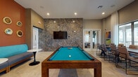 This thoughtfully designed single-floor amenity center offers a variety of well-appointed spaces for socializing, relaxing, and gathering. The main lounge is a standout area, anchored by a dramatic floor-to-ceiling stone accent wall with a sleek linear fireplace and a wall-mounted TV. A statement circular chandelier with exposed bulbs adds warmth and sophistication, while large windows dressed in flowing white curtains flood the space with natural light and provide views of the surrounding community. Adjacent to the lounge is a fully equipped kitchen featuring warm wood cabinetry, stainless steel appliances, a teal glazed brick backsplash, and an expansive white quartz island with bar seating — ideal for catered events or casual gatherings. A billiards room offers recreational appeal, complete with a pool table set against another striking stone-clad feature wall and a mounted TV. A dedicated coffee and beverage station with wood cabinetry serves as a welcoming touchpoint near the entrance. Multiple seating areas throughout include a vibrant lounge with teal velvet chairs and bold artwork, as well as a private dining room with a large communal table and garden views. Light tile flooring, curated artwork, and designer lighting fixtures carry a cohesive modern aesthetic throughout the entire space.