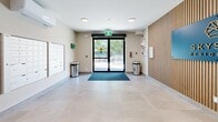 The lobby hallway at Bayside Residences offers a clean, contemporary first impression designed with both functionality and aesthetics in mind. Large-format beige ceramic floor tiles span the entire space, reflecting the recessed ceiling lighting and contributing to an open, airy atmosphere. A striking feature wall clad in vertical wood-slat paneling anchors one side of the lobby, displaying the Bayside Residences branding in a bold blue signage panel — immediately establishing the building's identity upon entry. Lush potted tropical plants are positioned throughout, adding warmth and a touch of greenery to the neutral palette. The main entrance consists of a full-height glazed door with a black frame, allowing natural light to filter into the space and offering a clear view of the outdoors. A recessed entry mat provides a practical transition point at the threshold. Along the opposite wall, a large bank of stainless steel resident mailboxes is neatly built in, accompanied by stainless steel recycling bins for convenience. A wall-mounted mini-split climate control unit ensures year-round comfort. Safety and building management elements, including an intercom panel, emergency signage, and a security camera, are thoughtfully integrated without disrupting the space's polished, modern character.
