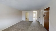 Located at 2 South Glenwood Avenue in Clifton Heights, Pennsylvania, this single-floor residence offers a straightforward and functional layout designed for comfortable everyday living. The main living area is generously proportioned, featuring wall-to-wall carpeting in a neutral tone that extends throughout the space, creating a cohesive and understated aesthetic. Crisp white walls and a clean white ceiling contribute to a bright, open atmosphere, while baseboard heating units ensure year-round comfort. An open passageway connects the main living area to an adjacent room, offering a natural flow between spaces. A hallway extends further into the unit, providing access to additional rooms and suggesting a well-organized floor plan. A closet with bi-fold doors is visible near the entry, offering convenient storage. Natural light filters in from windows at the end of the hallway, lending the interior a welcoming sense of brightness. The overall design is simple and practical, with neutral finishes throughout that provide a versatile backdrop suited to a variety of furnishing styles. The unit's single-level layout makes for easy navigation and accessibility throughout the home.