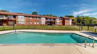 Located at 19 West Lakeshore Drive in Homewood, Alabama, this outdoor amenity area centers around a generously sized swimming pool with clear, blue-green water and a wide concrete surround that provides ample space for lounging and recreation. The pool deck is equipped with patio seating, including a table with an umbrella and additional chairs, making it a functional and inviting outdoor gathering space. The pool depth ranges from shallow to deep ends, as indicated by clearly marked depth markers along the pool's edge, with access provided via metal entry railings.

The pool area is enclosed by a sleek black iron fence that defines the space while maintaining open sightlines to the surrounding green lawns and mature deciduous trees, which offer natural shade and a lush, park-like atmosphere. Adjacent to the pool is a single-story utility or cabana-style structure with multiple compartment doors, providing convenient poolside storage or changing facilities.

The backdrop is defined by multi-story classic brick residential buildings featuring a warm red-brick exterior, a brown roof line, and large windows. The well-maintained grounds feature expansive grassy areas with established trees, contributing to a welcoming and serene outdoor environment. The space benefits from abundant natural light under open Alabama skies, creating a bright and pleasant atmosphere throughout.