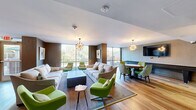 This thoughtfully designed single-floor amenity space offers a generous open layout spread across two connected zones, blending a sophisticated lounge area with a dedicated game and entertainment room. The interior is anchored by warm hardwood-style flooring and richly textured herringbone-patterned accent walls that lend the space a contemporary, upscale character throughout.

The main lounge area is furnished with plush neutral sofas and a bold collection of chartreuse green accent chairs, creating a vibrant mid-century modern aesthetic. Striking sputnik-style chandeliers serve as dramatic focal points overhead, while floor-to-ceiling glass windows and sliding doors flood the space with natural light and provide sweeping views of the surrounding landscaped outdoor area and patio.

A dining and bar seating zone features both a live-edge wood high-top table and sleek white counter-height surfaces, accommodating casual gatherings and social events. A wall-mounted television adds a media component to this area. Adjacent to it, the game room includes a full shuffleboard table, a round poker table surrounded by patterned upholstered chairs, and an additional large-screen television — ideal for entertaining.

Tasteful artwork adorns the walls, and the space connects seamlessly to an exterior patio with seating, extending the social experience outdoors. The overall ambiance is energetic yet refined, well-suited for community gatherings and leisure.