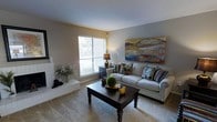 This two-floor space offers a comfortable and thoughtfully designed living environment. The living room serves as a welcoming focal point, featuring soft, neutral-toned walls that create a warm and inviting atmosphere. Wall-to-wall carpeting adds to the coziness underfoot, while a large window fitted with blinds allows natural light to filter into the room, providing views of the surrounding greenery outdoors.

A white brick fireplace anchors one corner of the living room, adding a classic architectural element and a sense of warmth to the space. The room comfortably accommodates a plush sofa accented with decorative pillows, paired with a dark wood coffee table that grounds the seating area. A floor lamp provides additional ambient lighting, complementing the natural light from the window. A bold, colorful landscape-inspired artwork displayed above the sofa adds a vibrant artistic touch to the otherwise understated décor.

The layout flows naturally across two floors, offering separation between living and other functional areas of the home. Overall, the space balances practicality with a relaxed, lived-in aesthetic suited for everyday comfort.