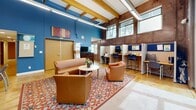 Located in Lakewood, Colorado, this single-floor community room offers a thoughtfully designed multipurpose space that balances comfort with functionality. The room's most striking architectural feature is its soaring ceiling, accented by exposed natural wood beams that add warmth and character to the otherwise modern industrial aesthetic. The walls blend rich wood paneling with bold blue and neutral tones, creating a dynamic yet inviting atmosphere.

The lounge area serves as the centerpiece of the space, anchored by a vibrant patterned area rug in red, cream, and blue. Cognac leather seating — including a full sofa and matching armchairs — is arranged around a round coffee table, offering a welcoming setting for conversation or relaxation. Expansive floor-to-ceiling glass sliding doors flood the room with natural light and provide direct views of the surrounding outdoor greenery and courtyard.

On one side of the room, a row of semi-private workstation cubicles with built-in desks and task seating provides a dedicated space for focused individual work. Additional seating and table arrangements along the perimeter further expand the room's versatility. Modern linear pendant lighting complements the natural light throughout. Together, these elements create a bright, functional, and welcoming environment well-suited for both collaborative and independent use.