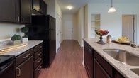 Located at 9417 Cumberland Dr in Fredericksburg, Virginia, this single-floor residence offers a cohesive and functional layout designed for comfortable everyday living. The galley-style kitchen is a standout feature, showcasing rich dark espresso cabinetry paired with granite-look countertops, a full suite of black appliances including a refrigerator, range, microwave, and dishwasher, and warm hardwood-style flooring underfoot. Track lighting and pendant lights illuminate the workspace, while a kitchen island with a double sink adds both utility and style. Built-in shelving near the entryway provides convenient storage and display space, and the kitchen flows naturally toward the main hallway and living areas. The bedroom is bright and inviting, finished with plush wall-to-wall carpeting, neutral walls, and a ceiling fan, with a window fitted with blinds that allows natural light and outdoor views. The bathroom continues the dark cabinetry theme with a vanity featuring a light countertop and integrated sink, a tub-shower combination with tiled surround, and hardwood-style tile flooring. Crown molding accents and white trim details throughout lend a polished finish to the space, creating a clean, consistent aesthetic from room to room.