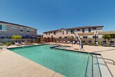 Located at 13025 W Thomas Rd in Avondale, Arizona, this outdoor community amenity area centers around a generously sized swimming pool with clear, blue-green water that reflects the abundant Arizona sunshine. The pool is complemented by an adjacent spa, providing residents with a range of aquatic relaxation options. An ADA-compliant pool lift is prominently positioned at the pool's edge, ensuring accessibility for all users.

The surrounding deck is finished in a neutral aggregate concrete surface and is well-appointed with multiple lounge chairs arranged for sunbathing and leisure. Shade structures, including sail-style canopies and freestanding umbrellas in navy blue, offer respite from the desert sun. Outdoor dining sets with umbrella-covered tables provide additional gathering space within the enclosure.

The entire area is enclosed by a black metal security fence with a gated entrance, offering both safety and privacy. Lush, young desert-adapted plantings are interspersed throughout the perimeter, adding greenery to the hardscape. The backdrop of the multi-story stucco residential buildings frames the space, consistent with the Southwestern architectural aesthetic of the surrounding community. Under the clear blue skies typical of Avondale, this pool area serves as a well-equipped and inviting outdoor retreat.