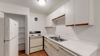 Located at 14417 Hellenic Dr in Tampa, Florida, this single-floor unit offers a practical and well-organized layout. The kitchen is designed for efficiency, featuring white cabinetry with wood-trimmed accents, a double-basin stainless steel sink, a coil-top electric range, a dishwasher, a full-size refrigerator, and light wood-style flooring. A wall-mounted ventilation fan and a dedicated pantry with built-in shelving provide added functionality. The open kitchen area transitions seamlessly into the adjoining living space, which features the same wood-style flooring and a clean, bright aesthetic with white walls. The living and bedroom areas are finished with wall-to-wall carpeting in a neutral tone and are painted in warm beige hues. Large windows fitted with vertical blinds allow natural light to filter in, offering views of the surrounding greenery. The bathroom is finished with white tile surrounding a full soaking tub and shower combo, complemented by wood-style flooring and brushed nickel fixtures. Additional storage is provided through a louvered bi-fold door closet with a full-width hanging rod. Textured ceilings are consistent throughout the unit.