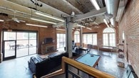 This single-floor space showcases a distinctive industrial character, blending raw architectural elements with functional amenities. The main open area is defined by exposed red brick walls, bare concrete ceilings with visible ductwork and conduit, and sturdy structural columns that reinforce the building's historic industrial roots. Ceiling fans and fluorescent strip lighting provide practical illumination throughout the expansive interior.

The lounge area is anchored by a brick fireplace with a mounted flat-screen television, surrounded by leather seating arranged on a patterned area rug. A full-size pool table occupies a central position in the space, adding a recreational focal point. Arched windows and large glass-paned doors flood the interior with natural light and provide views of an outdoor patio area.

A corridor connects the main space to additional rooms, featuring polished concrete floors, blue accent walls, exposed brick, and a wood-railed staircase with metal balusters. Off this hallway, a kitchen area is outfitted with white cabinetry, a stainless steel refrigerator, a gas range, and dark countertops. The overall ambiance is bold and urban, combining the character of a repurposed historic building with versatile, well-appointed interior spaces suitable for a variety of uses.