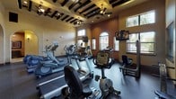 Located at 650 Leora Lane in Lewisville, TX, this fitness center presents a thoughtfully designed single-floor facility that blends functionality with a warm Mediterranean-inspired aesthetic. The space is defined by its distinctive exposed dark wood beam ceilings, accented by ceiling fans and recessed lighting that create a comfortable workout environment throughout.

The cardio zone is generously appointed with treadmills, elliptical trainers, stationary bikes, and stair climbers, all positioned beneath large windows fitted with horizontal blinds that allow natural light to filter in while offering outdoor views. Wall-mounted televisions are strategically placed for convenience during cardio sessions.

A dedicated strength training area features a variety of cable machines, weight benches, and a full dumbbell rack accompanied by wall-to-wall mirrors, offering ample space for both machine-based and free weight exercises. The rubber flooring throughout provides durability and safety underfoot.

Elegant arched colonnades connect the workout zones to a tiled corridor, which leads to restroom facilities and a dual-level water fountain station for easy hydration. The warm earth-tone plaster walls, decorative wall sconces, and framed artwork in the connecting hallway add a refined character that distinguishes this fitness center from standard facilities.