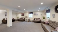 This single-floor space is thoughtfully designed to accommodate group living and communal activities, offering a warm and welcoming atmosphere throughout. The expansive lounge area serves as the centerpiece of the space, featuring plush dark brown leather sofas accented with mauve and purple throw pillows, arranged across a soft neutral-toned carpet. Recessed lighting illuminates the room evenly, while multiple windows dressed with decorative valances allow natural light to fill the space. Wall art and potted greenery add tasteful decorative touches, contributing to a calm and comfortable ambiance. A wall-mounted flat-screen television is also present for residents' use. Adjacent to the lounge, the dining area offers generous seating capacity with multiple high-top tables crafted from rich dark stone-look surfaces, paired with black leather chairs. Natural light enters through several windows, and a mounted television is available in this area as well. The kitchen is fully equipped with warm honey-toned wood cabinetry, dark granite-style countertops with a matching backsplash, stainless steel appliances including a range, over-the-range microwave, and refrigerator, along with a double sink. A pass-through window connects the kitchen to the dining area, supporting efficient meal service. Light wood-style flooring runs through the kitchen and dining spaces, tying the functional areas together cohesively.