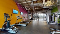 Located at 500 W Broad St in Columbus, Ohio, this single-floor fitness center delivers a dynamic and energizing workout environment designed to motivate and perform. The space is immediately distinguished by its bold, color-blocked walls in vivid shades of magenta, orange, and green, creating a visually stimulating atmosphere that sets it apart from a typical gym setting.

The exposed concrete ceiling with visible ductwork and industrial-style piping lends an urban, loft-like aesthetic throughout. Durable rubber flooring covers the entire space, providing both safety and functionality for a range of exercise activities.

The cardio zone is well-appointed with a row of Star Trac treadmills, an elliptical, and stationary bikes, each equipped with personal screens for entertainment during workouts. Mounted flat-screen televisions on the accent walls provide additional viewing options. Large floor-to-ceiling glass garage doors line one wall, flooding the space with natural light and offering views of the surrounding streetscape, creating an open, airy feel.

The strength training area features a cable machine, a full rack of dumbbells, a weight bench, and ample open floor space for functional training. Full-length mirrors reflect the colorful interior, enhancing the sense of spaciousness. This well-equipped facility combines industrial design with energetic aesthetics to create an inviting and comprehensive fitness destination.