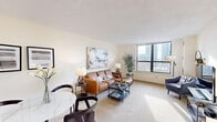 This single-floor residence offers a thoughtfully designed layout that balances comfort and functionality throughout its interconnected living spaces. The open-concept living and dining area is bright and welcoming, anchored by large floor-to-ceiling windows that frame impressive urban skyline views and flood the carpeted interior with natural light. The living area provides generous space for seating arrangements, while a dedicated dining zone accommodates a full table set, complemented by a built-in pass-through window connecting directly to the kitchen. A compact workspace nook adds further versatility to this shared living space. The galley-style kitchen is efficiently organized with warm wood cabinetry lining both sides, light countertops, tile flooring, and full appliances including a range, oven, and refrigerator. The bedroom is a serene retreat, offering plush carpeting, a large closet with paneled wooden doors, and additional city views through a generous window. The full bathroom features tile flooring, a wood-toned vanity with an integrated sink, a large wall mirror with Hollywood-style lighting, and ample counter space. Throughout the residence, neutral tones and soft textures create a cohesive and calm ambiance, well-suited for urban living.